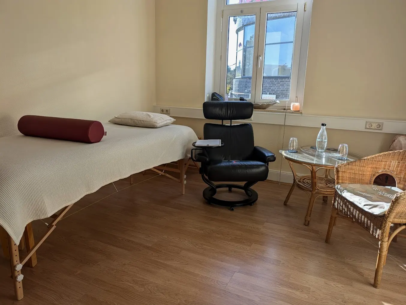 Praxisraum von Seelenessenz in Sankt Augustin: Liege mit heller Decke, Sessel und Rattan-Sitzgruppe am Fenster
